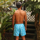 Man wearing blue swim shorts with a floral pattern standing outdoors among greenery.