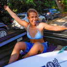 Woman in a blue bikini sitting on a surfboard in a car's open trunk with cars and greenery in the background.