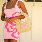 Person wearing a pink dress with a floral design, holding a straw bag with a pink flower.
